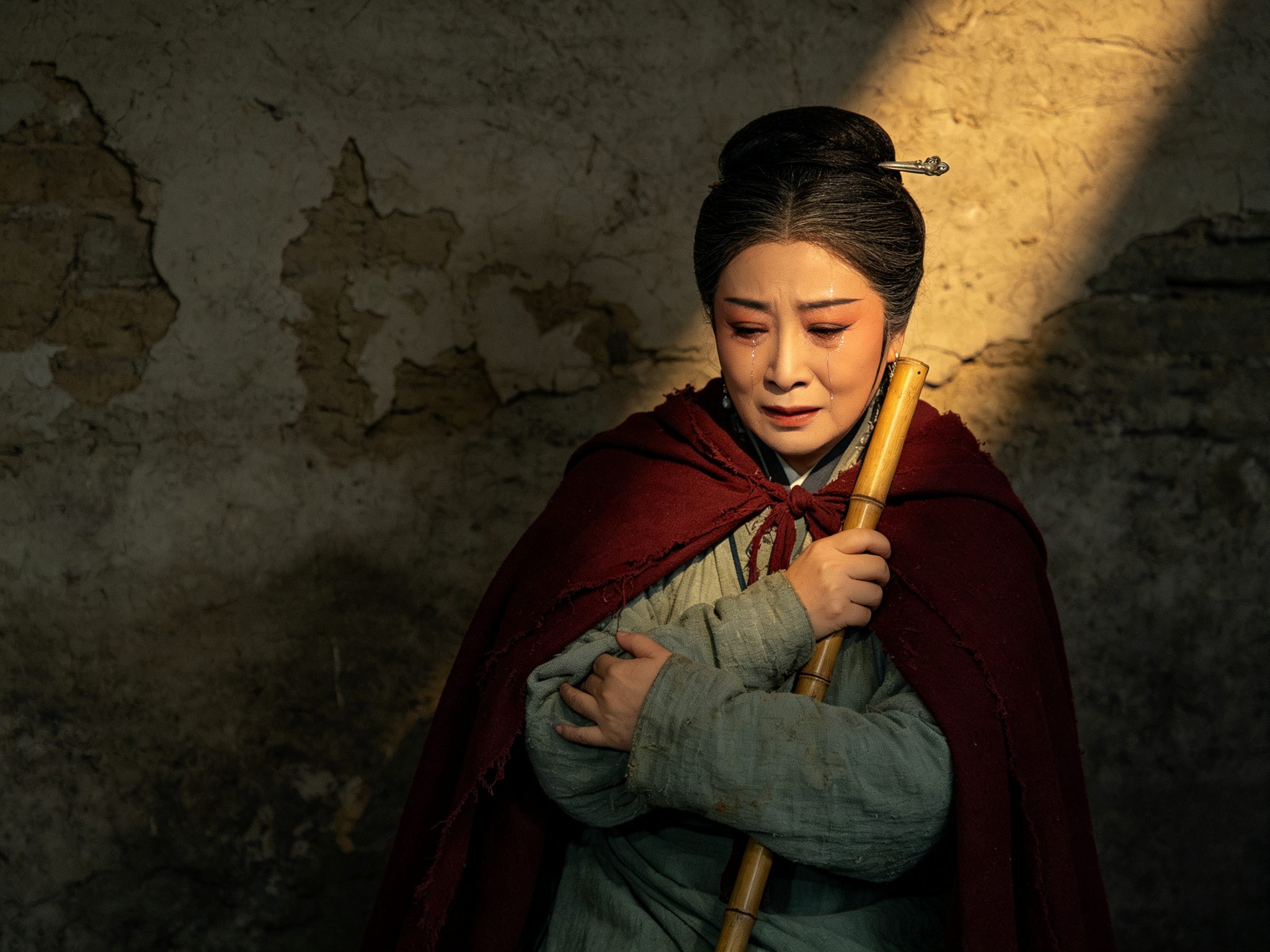 秦腔《狸猫换太子》正旦李宸妃舞台造型，中年女性荆钗布裙、身披深红旧斗篷、手扶竹杖、眼角挂泪面容凄苦，斑驳寒窑土墙前一束追光打下