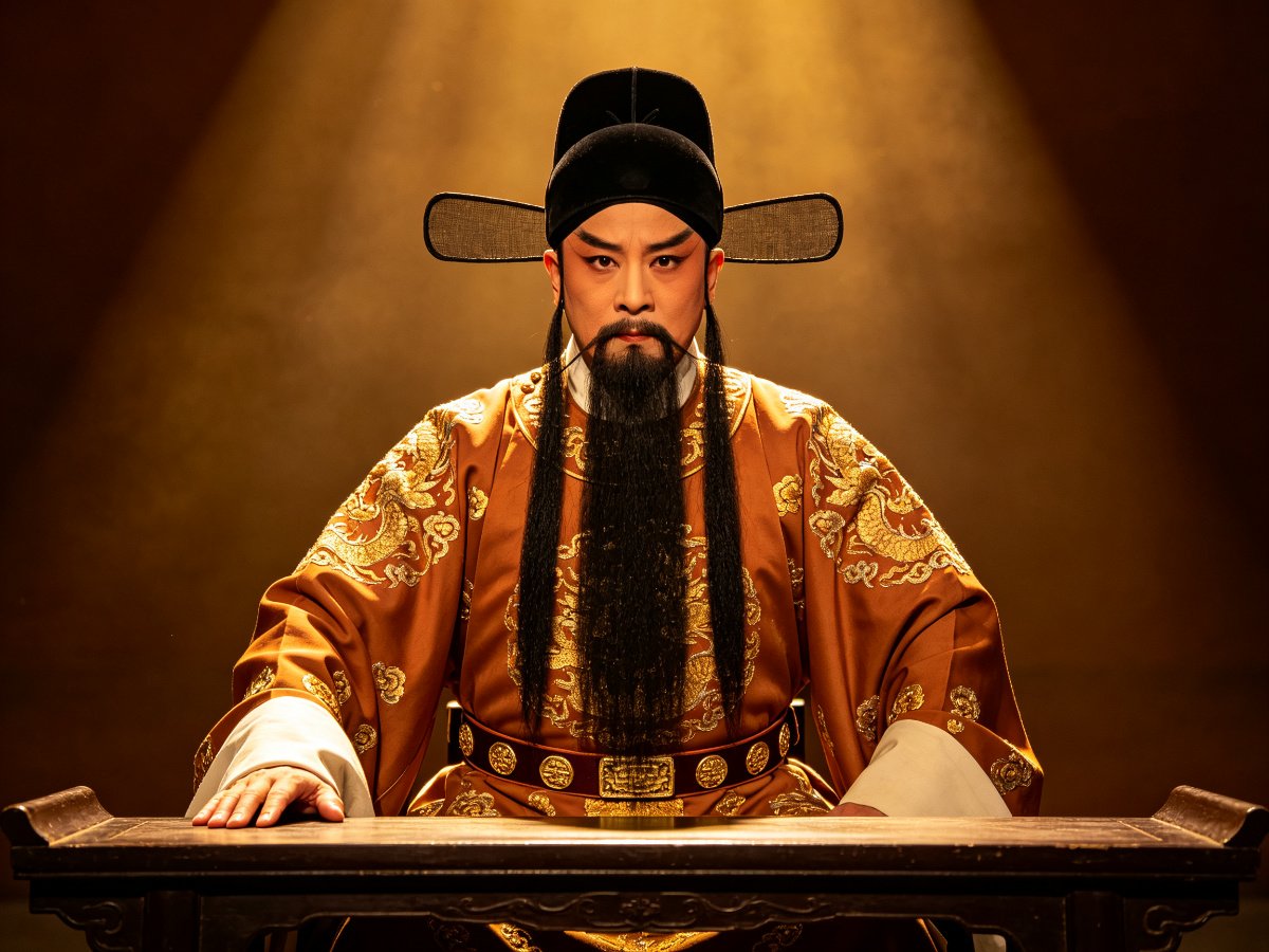 豫剧老生徐达形象，挂黑三髯、头戴乌纱帽、身穿赭金色团花官袍，端坐公案之后、一手拍案、目光如炬